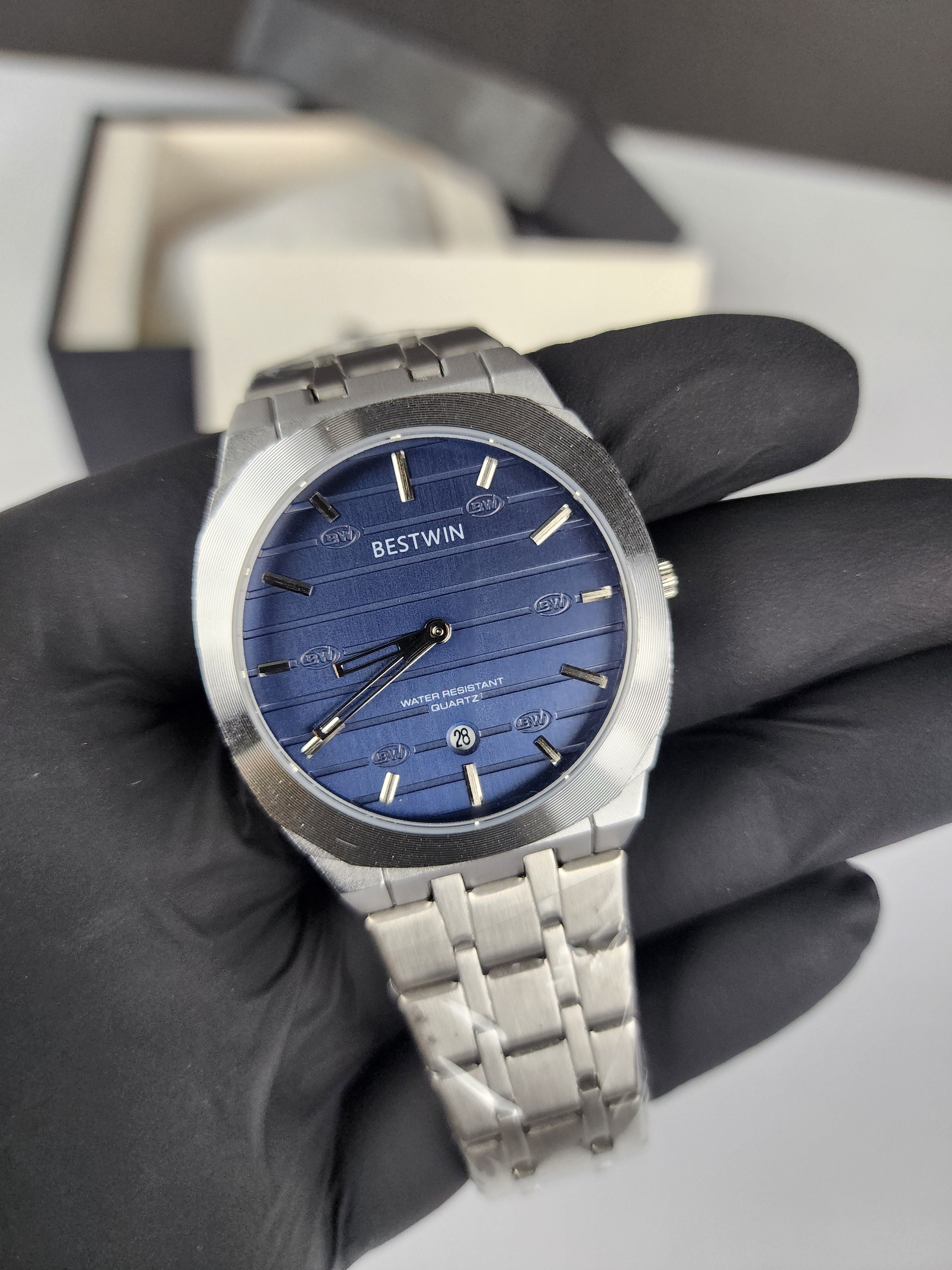 Silver watch with blue face held by a gloved hand against a neutral background